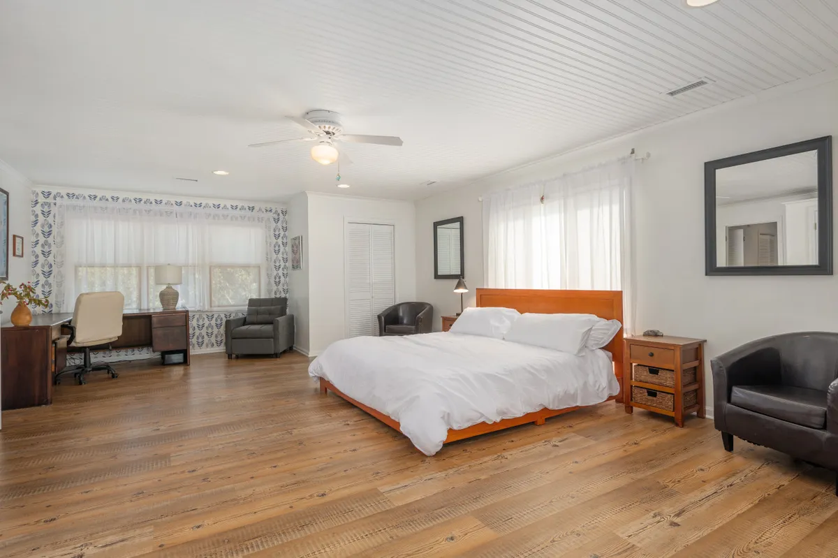 A serene bedroom featuring a plush bed, a desk positioned near a sunlit window, and comfortable armchairs. Natural light streams in, illuminating the workspace with views outside, creating an ideal spot for rest or productivity.