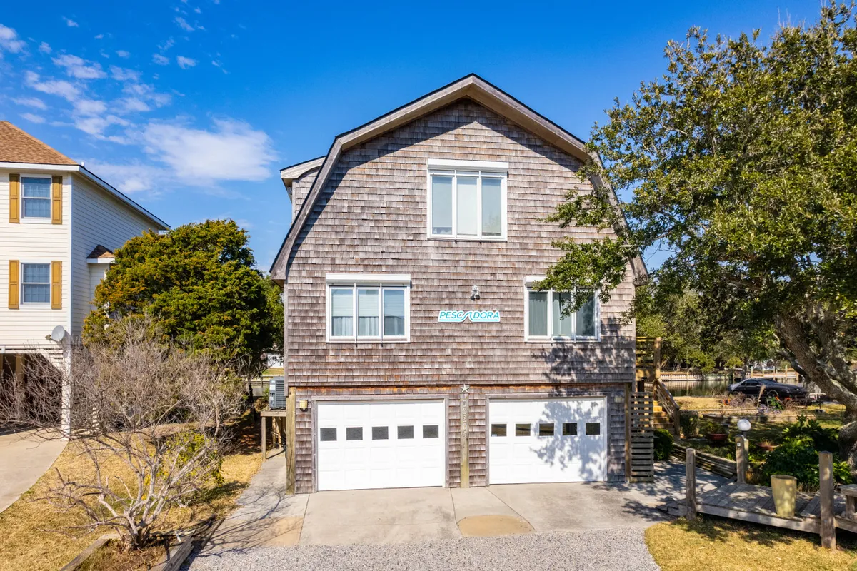 The exterior of the home is shown with large windows, wooden siding, and two prominent garage doors, surrounded by lush greenery. The double garage is easily visible, as is the spacious driveway area suitable for multiple vehicles.