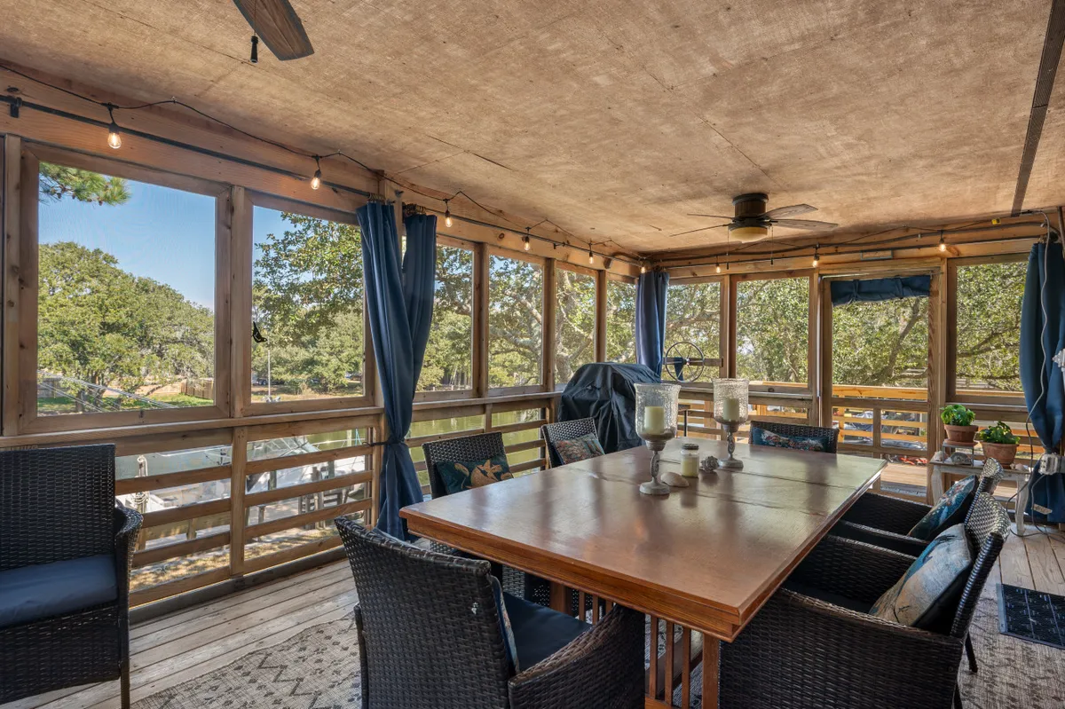 A spacious, sunlit dining area features a large wooden table surrounded by wicker chairs, with expansive windows offering beautiful views of the waterfront. A potted plant sits at the center, enhancing the comfortable and relaxed mood of the space.