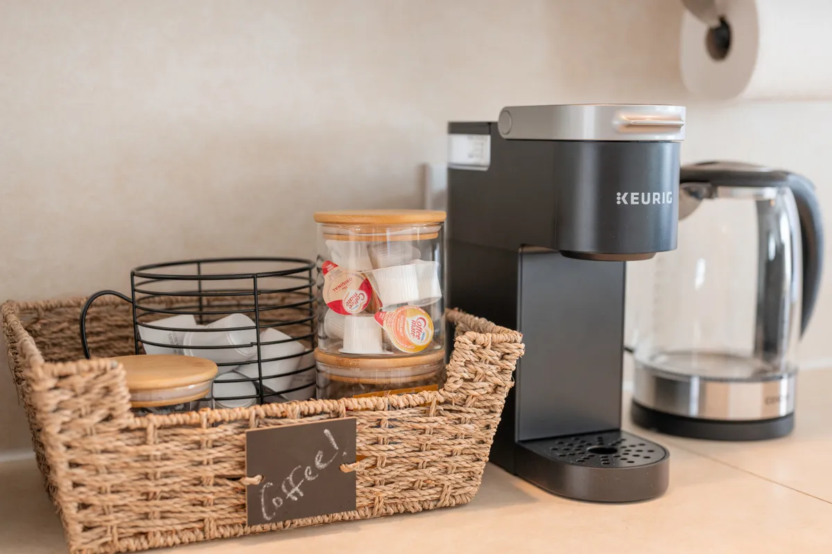 A cozy coffee station is set up with a Keurig coffee maker, a basket of coffee supplies, and a decorative jar, suggesting a warm, inviting spot for morning routines or socializing over a hot beverage.