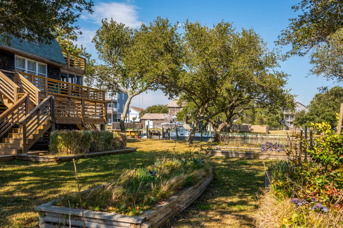 An exterior deck overlooking lush greenery and a water view, with garden beds nearby. The deck invites relaxation, outdoor dining, or quiet time with a pet, and the setting feels peaceful and open to nature.