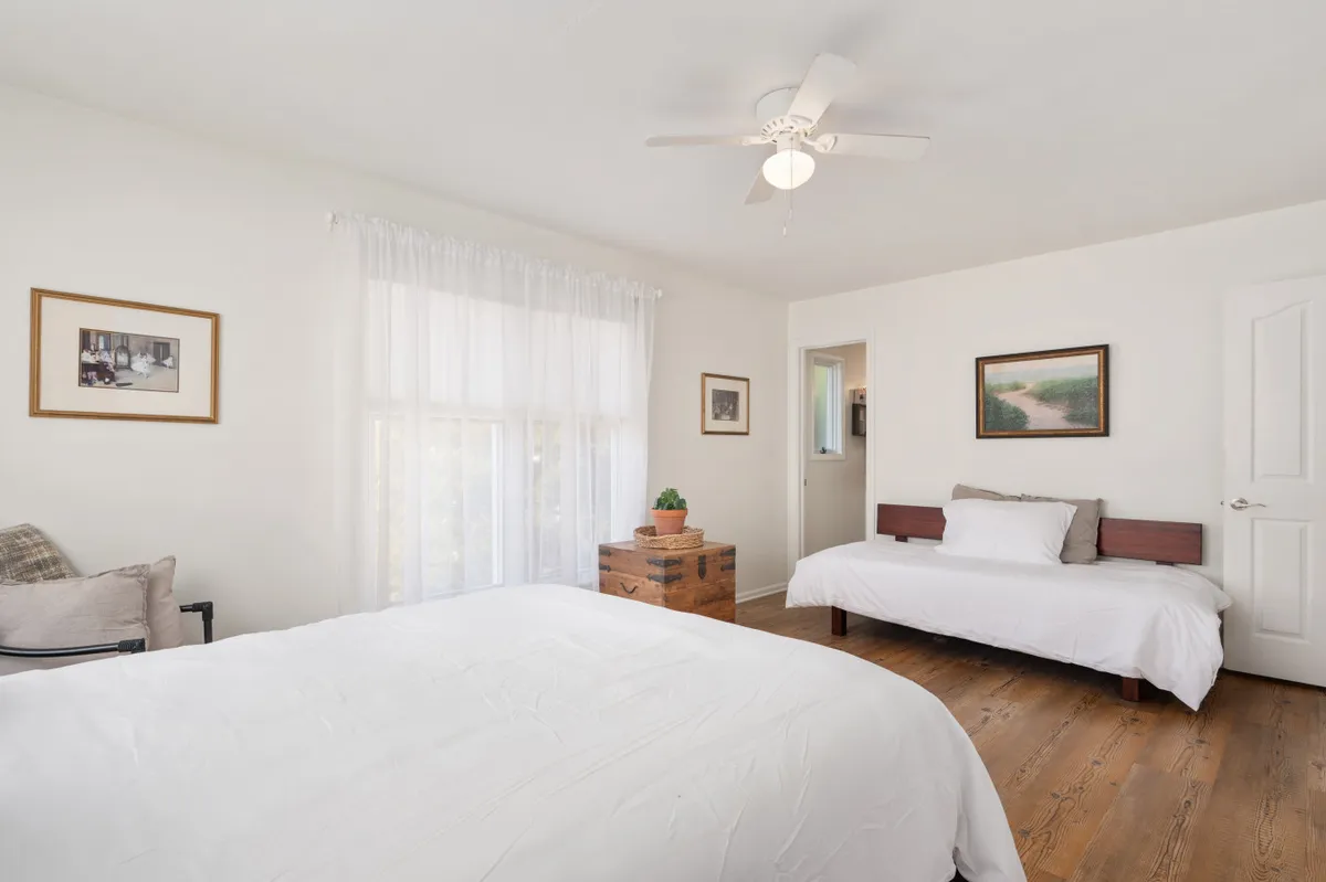 A cozy bedroom with two beds, a wooden chest, and large window draped in curtains. Soft natural light enters the room, creating a calm atmosphere suited for resting or evening chats.